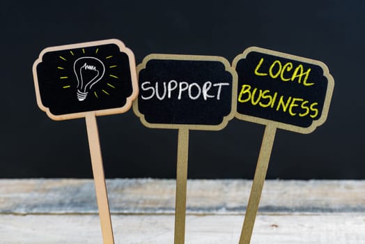 Concept message SUPPORT LOCAL BUSINESS and light bulb as symbol for idea written with chalk on wooden mini blackboard labels, defocused chalkboard and wood table in background