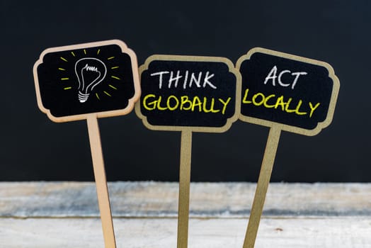 Concept message THINK GLOBALLY ACT LOCALLY and light bulb as symbol for idea written with chalk on wooden mini blackboard labels, defocused chalkboard and wood table in background