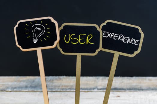 Concept message USER EXPERIENCE and light bulb as symbol for idea written with chalk on wooden mini blackboard labels, defocused chalkboard and wood table in background