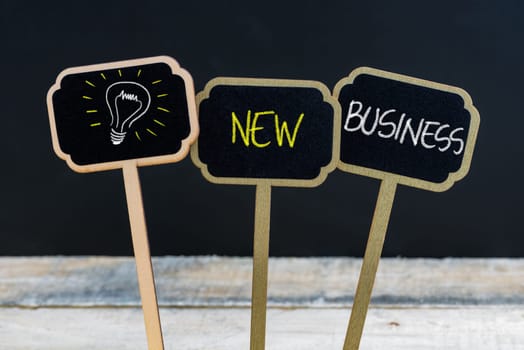 Concept message NEW BUSINESS and light bulb as symbol for idea written with chalk on wooden mini blackboard labels, defocused chalkboard and wood table in background