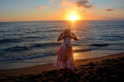 Girl in a long dress,holds a hat on her head with both hands and stands on the sea beach at sunset,with a view of the sun