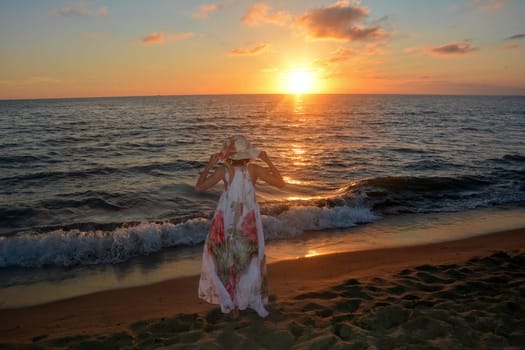 Girl in a long dress,holds a hat on her head with both hands and stands on the sea beach at sunset,with a view of the sun