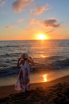 Girl in a long dress,holds a hat on her head with both hands and stands on the sea beach at sunset,with a view of the sun