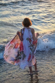 Girl from behind stands in the water on the beach,in the light of the sunset and holds up with hands long dress