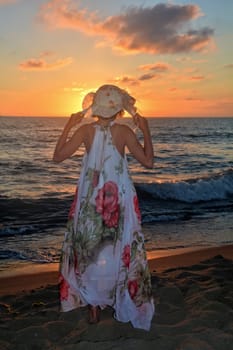 Girl in a long dress and holds hat on her head with both hands and stands on the sea beach at sunset,with a view to the sun,head covers the sun
