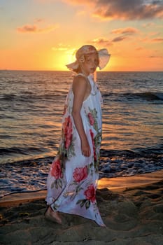 Girl in a long dress and hat on her head stands on the sea beach at sunset,looking forward