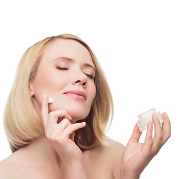 Beautiful middle aged woman with smooth skin and short blond hair applying moisturizing cream on face. Beauty shot. Isolated over white background. Copy space.