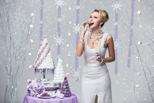 Beautiful young happy bride in felted gown eating wedding macaroon french style cake over snowy background and holding champagne glass