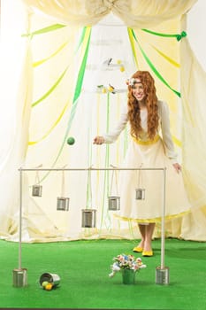 Beautiful young woman in yellow dress playing game, throwing ball into aluminium cans. Vertical composition.