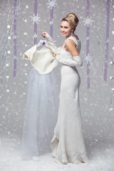 Beautiful happy young bride in felted winter dress holding veil cape on decorated hanger over snowy background