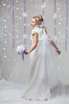 Portrait of beautiful young woman in long warm gown for winter wedding with purple flower bouquet. Over snowy background