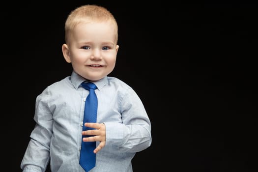 Cute baby boy in shirt, vest holding his blue tie.  Isolated over black background. Copy cpace.