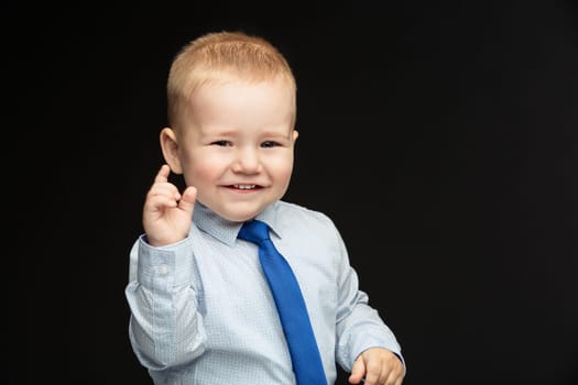 Cute baby boy in shirt, vest and tie showing something small. Over black background. Copy space.