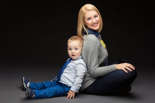 Beautiful mother and baby son sitting floor back to back. Happy people. Over black background.