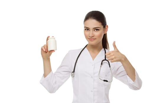 Beautiful young woman dressed as doctor holding container with pills and showing thumb up. Isolated over white background