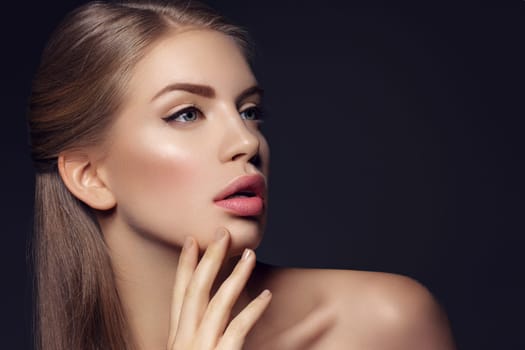 Beautiful young woman with blond hair and nude pink lips touching face. Over black background. Closeup beauty shot.
