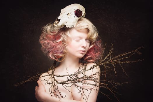 Portrait of beautiful young woman with animal scull on head and branches around her shoulders over dark background