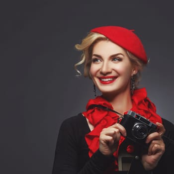 Beautiful young woman in red beret holding retro photo camera. Happy expression. Red lips. Over black background. Copy space.