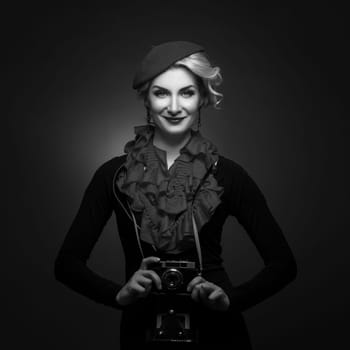 Beautiful young woman in beret holding retro photo camera. Happy expression. Monochrome. Over black background. Copy space.