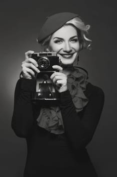 Beautiful young woman in red beret holding retro photo camera. Happy expression. Red lips. Over black background. Copy space.