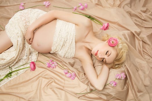 Beautiful young pregnant woman in lace fabric dreaming with flowers lying around