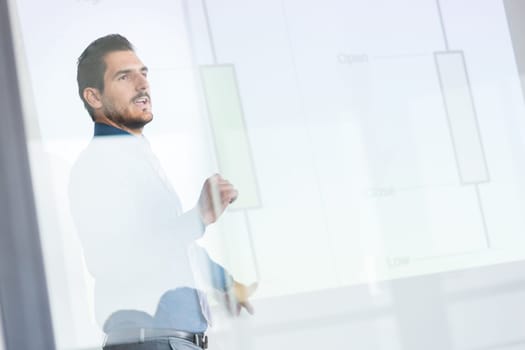Business man making a presentation in front of whiteboard. Business executive delivering a presentation to his colleagues during meeting or in-house business training. View through glass.