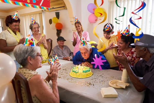 Group of old friends and family celebrating senior man 80 birthday party in retirement home. Happy elderly people having fun. Grandfather blowing candles on cake.