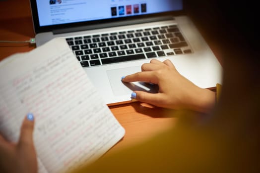 Young woman clicking on trackpad of laptop computer, doing a web search for homework. Female student studying at night and using pc at home. Over the shoulder shot