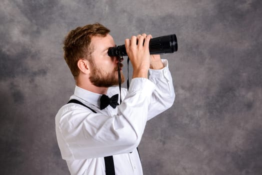 young bearded man in white shirt and bow tie looking through a binoculars