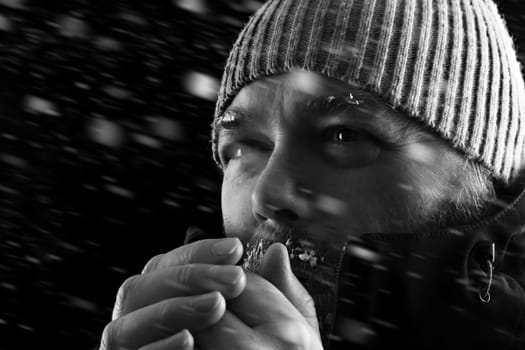 Freezing cold man standing in a snow storm blizzard trying to keep warm. Wearing a beanie hat and winter coat with frost and ice on his beard and eyebrows. Black and white. Looking to the left.
