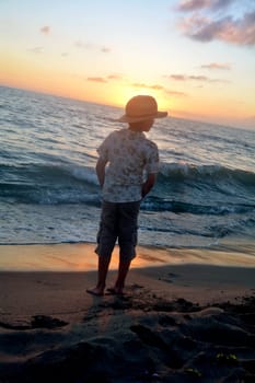 Little boy stands on the beach at sunset,hands in his pockets and looks to the side,head covers the sun