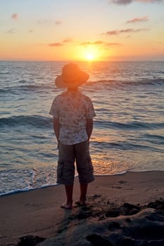 Little boy stands on the beach at sunset,hands in his pockets and looks at the sun