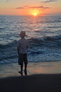 Little boy stands on the beach at sunset,hands in his pockets and looks at the horizon