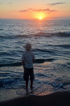 Little boy stands on the beach at sunset,hands in his pockets and looks at the horizon