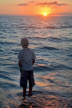Little boy stands in the waves on the beach at sunset,hands in his pockets and looks into the water