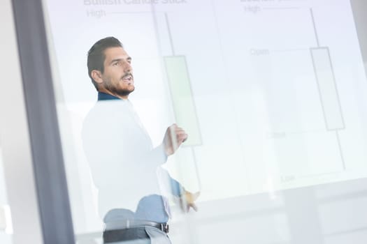 Business man making a presentation in front of whiteboard. Business executive delivering a presentation to his colleagues during meeting or in-house business training. View through glass.