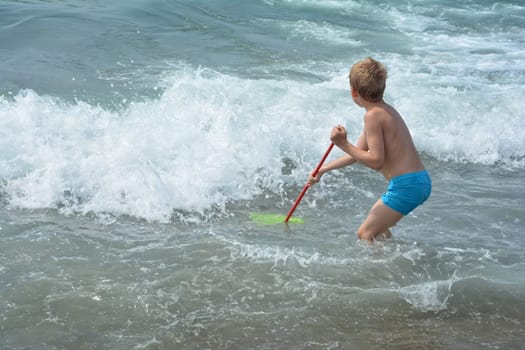 Little blond boy with blue swimming trunks stands in the water with a net and waits for the sea wave