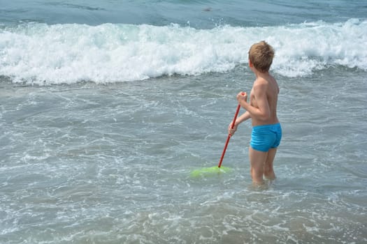 Little blond boy with blue swimming trunks stands in the water with a net and waits for the sea wave