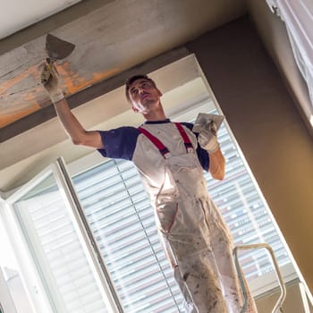 Construction worker wearing worker overall with wall plastering tools renovating apartment house. Plasterer renovating indoor walls and ceilings with float and plaster. Construction finishing works.