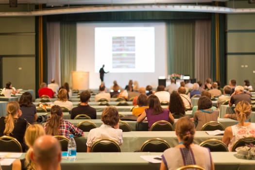 Business and entrepreneurship symposium. Speaker giving a talk at business meeting. Audience in the conference hall. Rear view of unrecognized participant in audience.