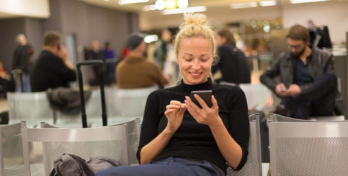 Casual blond young woman using her mobile phone while waiting to board a plane at airport departure gates.