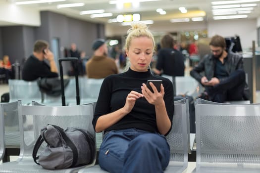 Casual blond young woman using her mobile phone while waiting to board a plane at airport departure gates.