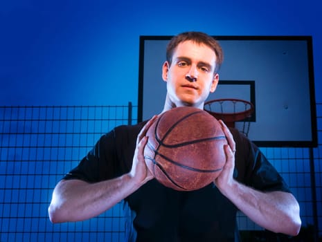 Frontal upper body view of a young man with basketball in front of a blue background at night with basketball hoop.