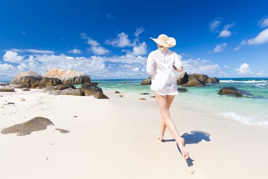 Happy woman wearing white loose tunic over bikini and beach hat, enjoying amazing white sandy beach on Mahe Island, Seychelles. Summer vacations on picture perfect tropical beach concept.