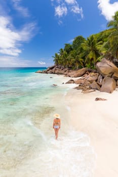 Woman wearing retro striped bikini and beach hat, enjoying amazing view on Anse Patates beach on La Digue Island, Seychelles. Summer vacations on picture perfect tropical beach concept.
