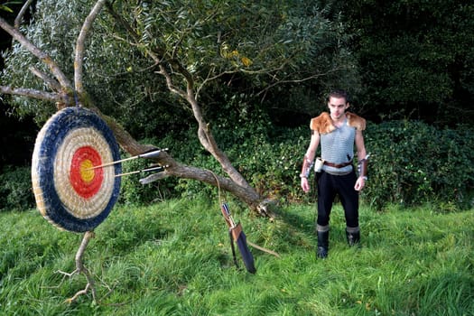 Medieval man stands in nature. Arrow,bow and quiver stands next to it and target made of straw with arrows hangs on the branch on the left side