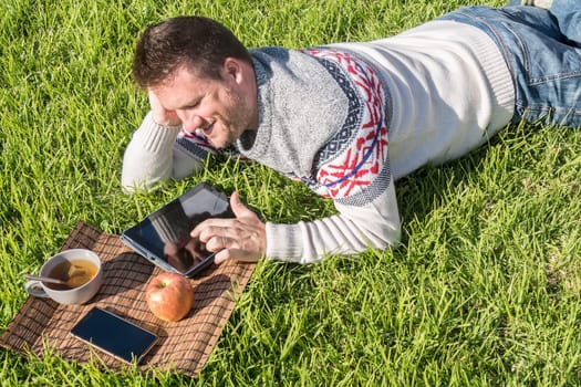 Man having breakfast lying on the grass in the garden with technology