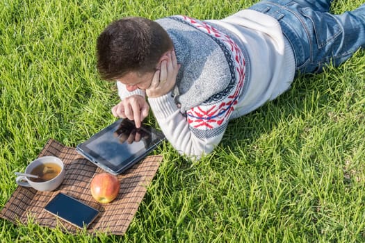 Man having breakfast lying on the grass in the garden with technology