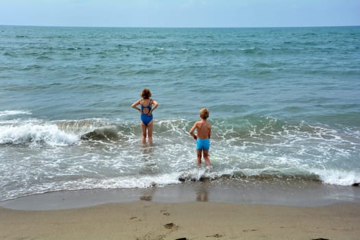 Boys and girls with blue swimwear stand in the water on the beach and look at the sea