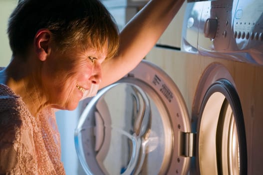 Head-and-shoulder view of a mature woman squatting in front of the open washing machine door and looking into the washing drum.